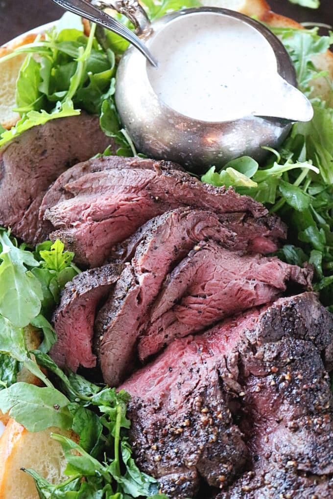 A close up shot of sliced roasted beef tenderloin sitting on top of a bed of mixed greens. Also nestled in the greens is a gravy boat of horseradish dressing. 