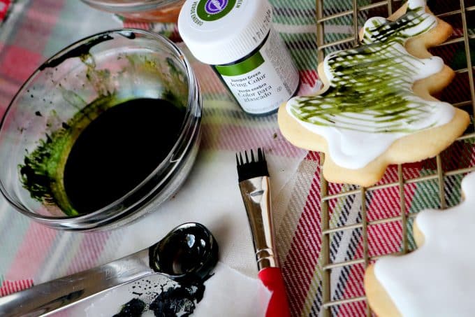 A glass bowl of green diluted icing, a teaspoon, paintbrush, jar of green gel food coloring and a iced Christmas tree cooking all sitting on top of a plaid tablecloth.  
