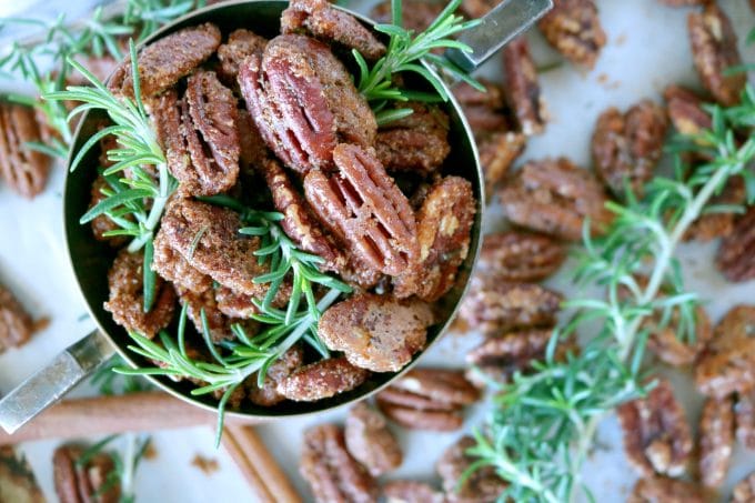 An overhead shot of candied pecans with fresh rosemary in a serving bowl. Scattered around the bowl are more pecans and fresh rosemary sprigs. 