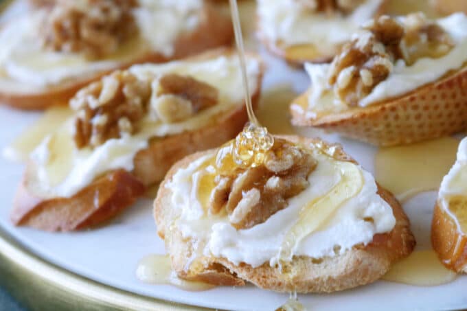 Honey goat cheese toasts on a white serving plate that is being drizzled with fresh honey on one of the toasts. 