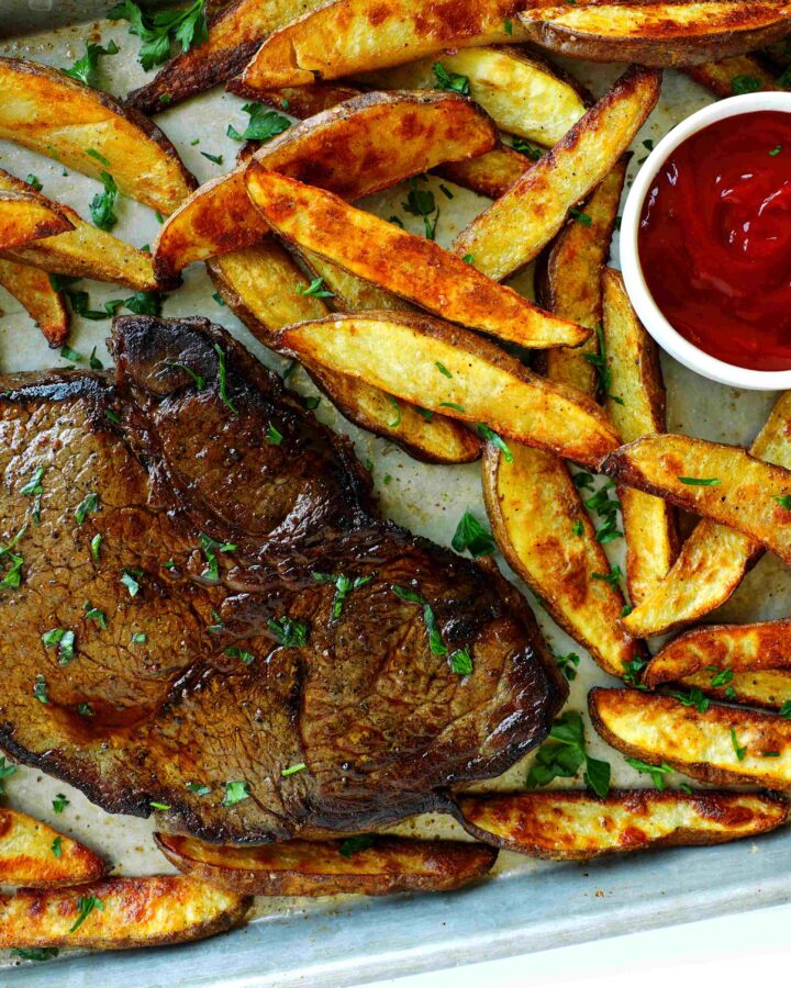 Ribeye steak served with crispy homemade steak fries and ketchup on a sheet pan, garnished with parsley.