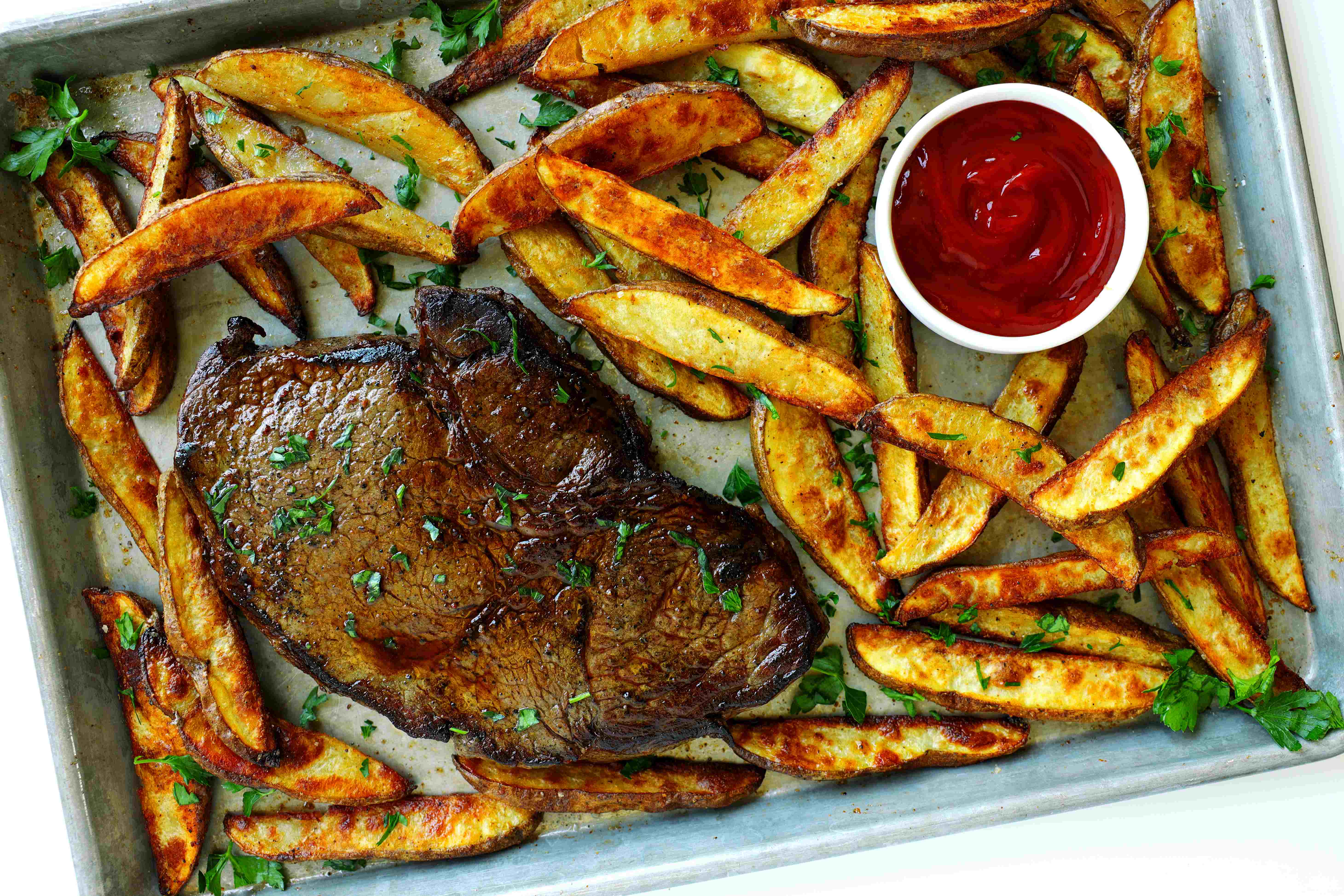 Ribeye steak served with crispy homemade steak fries and ketchup on a sheet pan, garnished with parsley.