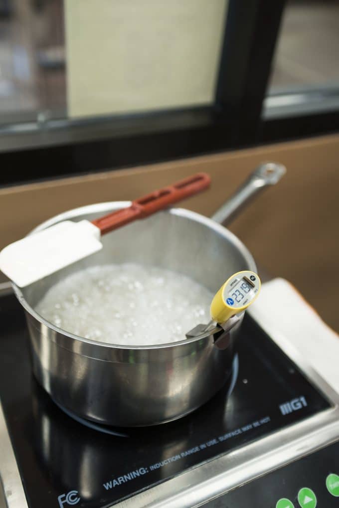 A picture of a pot that is boiling sugar. A spatula is lying flat across the pan along with a thermometer sitting inside the saucepan. 
