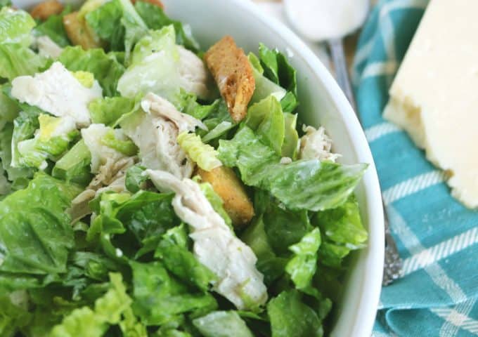 Shredded chicken sitting on a bed of romaine lettuce with a couple of croutons sprinkled in. The salad is in a white bowl and lying next to it is a teal and white napkin along with a block of parmesan cheese. 