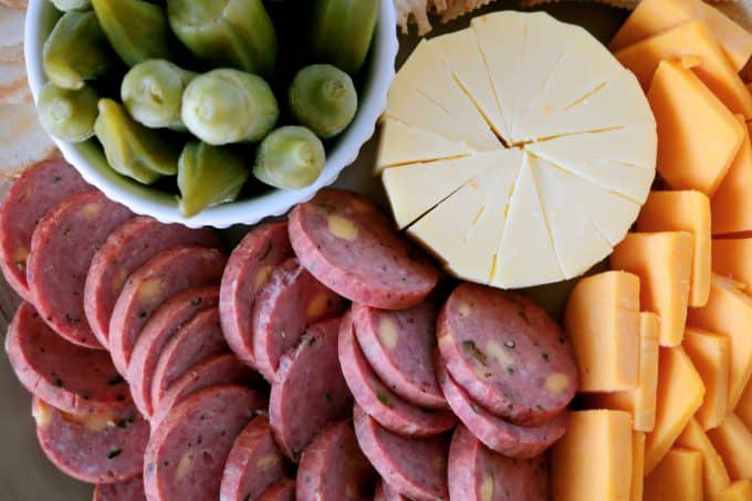 An overhead shot of summer sausage slices, smoked gouda, sliced yellow cheddar and a small white bowl of pickled okra. 