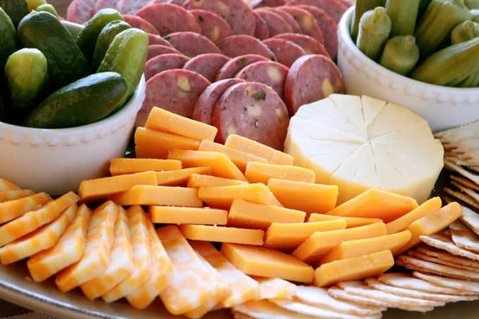 A close up shot of a cheese platter featuring colby jack, cheddar and gouda cheeses. In the back of the platter are slices of summer sausage. There are also two small white bowls sitting on the platter full of pickled okra and pickles. 