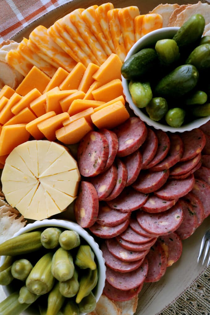 An overhead shot of smoked gouda, round sliced summer sausage, yellow cheddar and colby jack cheese. On each side of the platter are white bowls of pickled okra and pickles. 