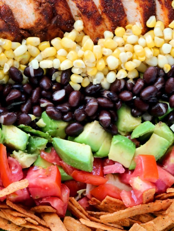 A close up shot of layered southwest chopped chicken salad ingredients. Starting at the bottom and going up are: tortilla strips, diced tomato, diced avocado, black beans, corn, sliced chicken. 