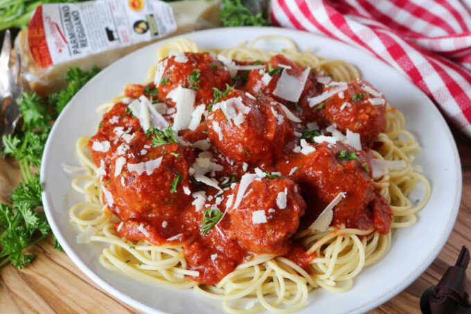 Spaghetti and Meatballs served in a white shallow bowl. The bowl is sitting on top of a wood cutting board surrounded by fresh parsley, a block of cheese, and a red and white checkered napkin. 