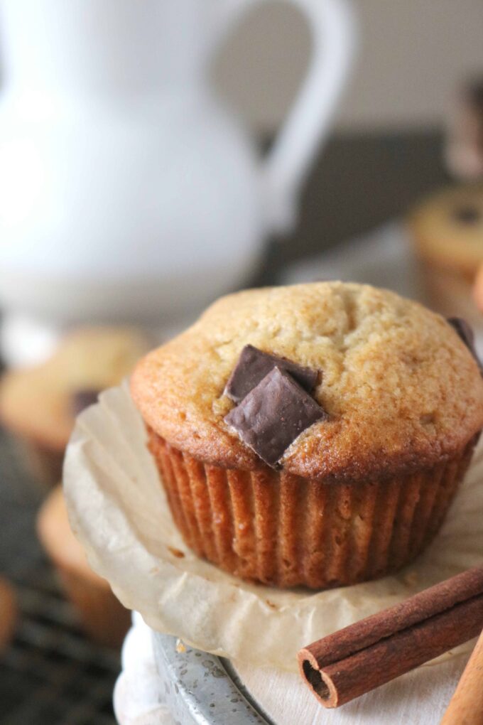 A close up shot of bakery style chocolate chunk muffins that has been unwrapped from the muffin wrapper with a cinnamon stick lying next to the muffin. 
