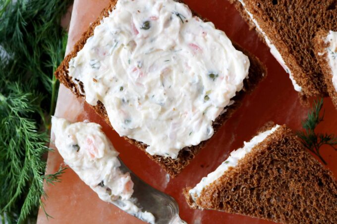An overhead shot of an open faced piece of pumpernickel bread with lox spread on top. Lying next to that are more sandwiches and a butter knife with a dollop of lox spread on it. 