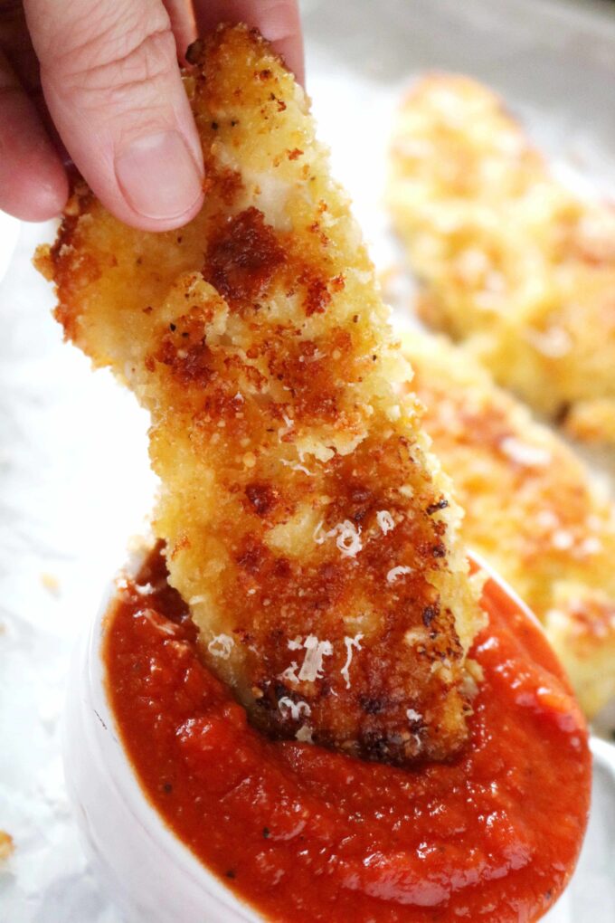 A hand dipping a parmesan crusted chicken tender into a bowl of marina sauce. 