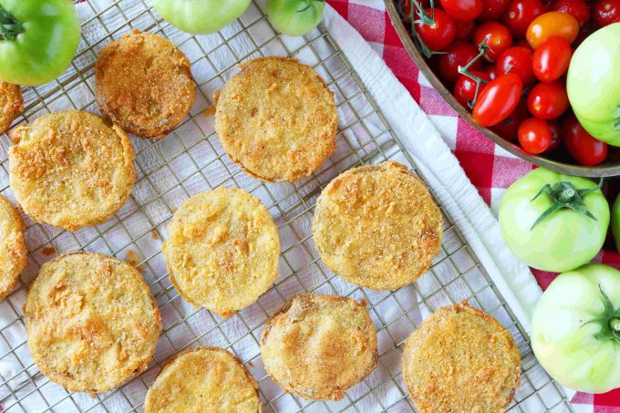 An overhead shot of easy fried green tomatoes placed on a wire rack. Next to the cooling rack are whole green and cherry tomatoes. 