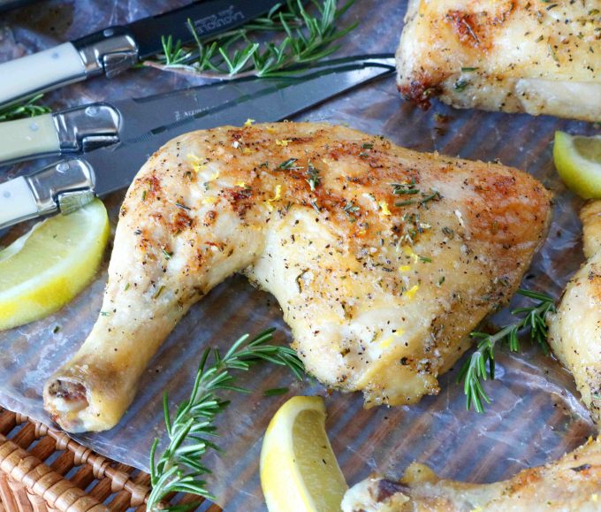 A baked chicken leg quarter on a tray surrounded by bundle of knives, lemons, and rosemary sprigs.