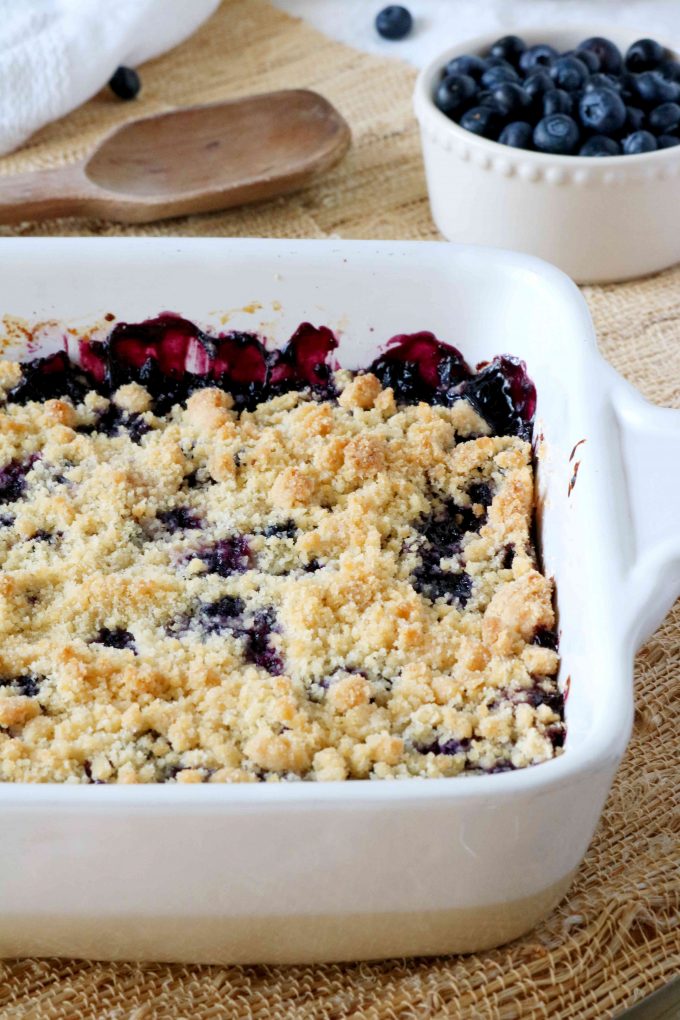 Half of a white casserole dish with baked blueberry bars. In the background is a wooden spoon and a bowl of fresh blueberries. 