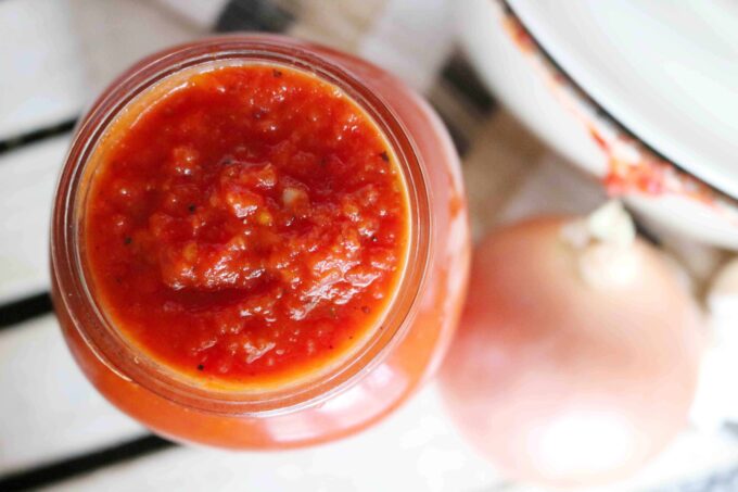 An overhead shot of italian marinara sauce in a clear glass har. Sitting next to the jar is a whole onion. 