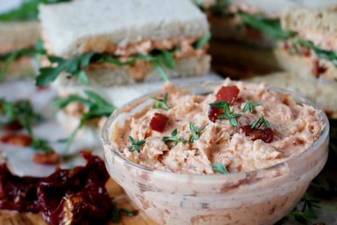 The image is focused on a clear glass bowl of sun-dried tomato spread. In the background there are prepared sandwiches. 