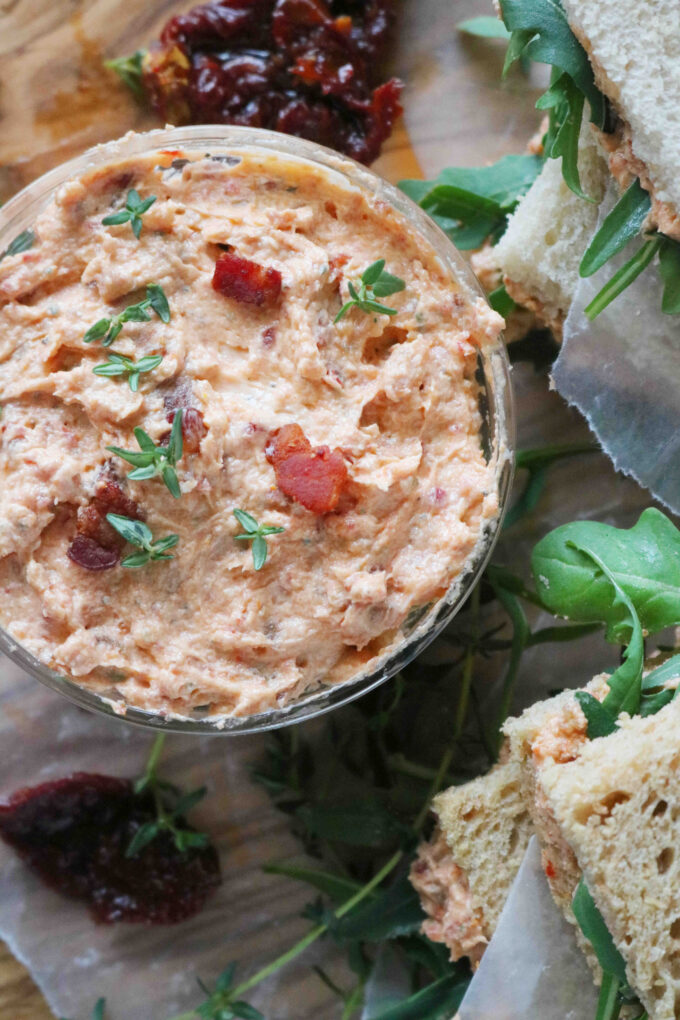 An overhead shot of sun-dried tomato spread in a small bowl. Surrounding the bowl are fresh herbs, cranberries and a couple of prepared sandwiches. 