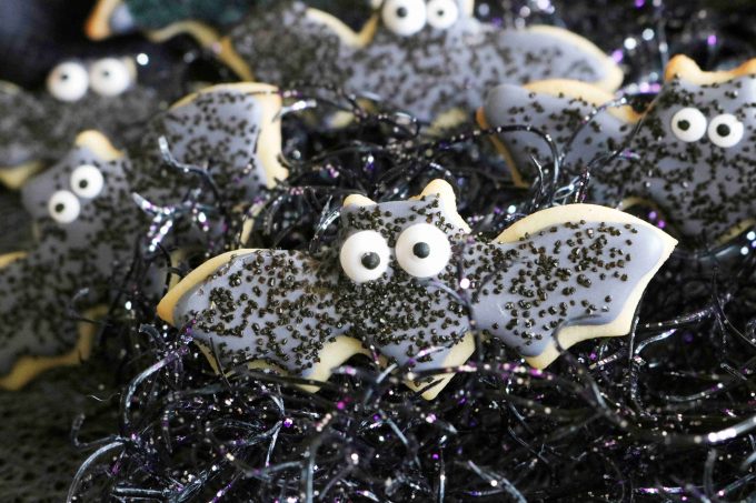 Halloween sugar cookie shaped like bats. The icing is black topped with black sprinkles along with two eye sprinkles. 
