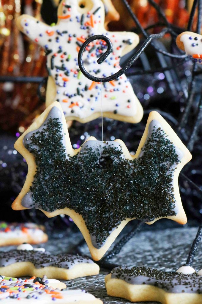 A bat shaped sugar cookie hanging from a black wire stand. The cookie is topped with black icing and sprinkles. In the background are more decorated halloween cookies. 