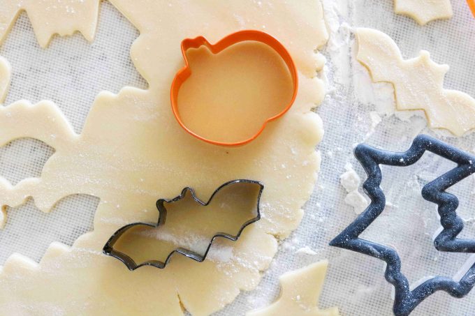 An overhead shot of a orange pumpkin cookie cutter, black bat and larger black bat cookie cutter on top of sugar cookie dough. 