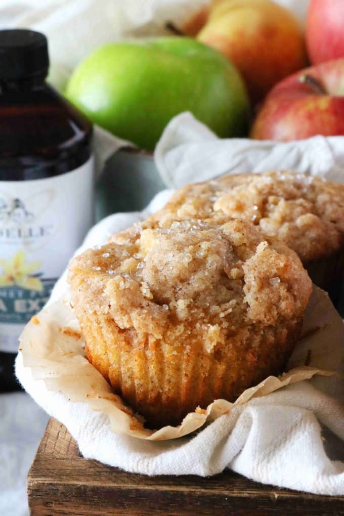 Two Apple Crumble Muffins sitting on top of white napkin with apples and vanilla extra in the background. 