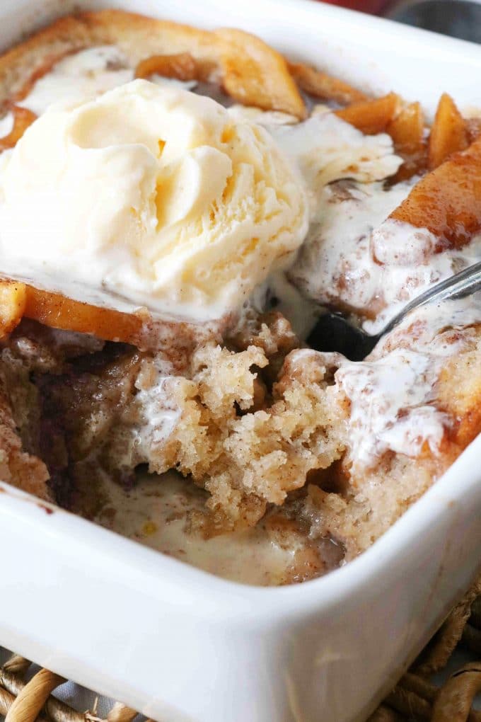 A close up shot of apple pudding cake with a piece missing from the white baking dish. In the center of the dish is a scoop of vanilla ice cream. 