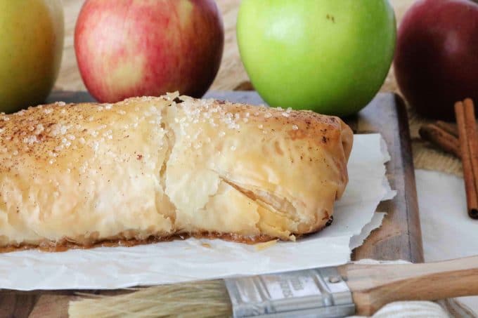 Apple Strudel that is placed on parchment paper with whole red and green apples sitting behind the cooked pastry. 