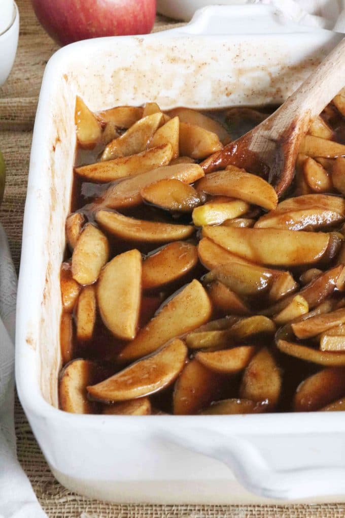 Baked cinnamon apples being baked in a white baking dish with a wood spoon resting on the side of it. 