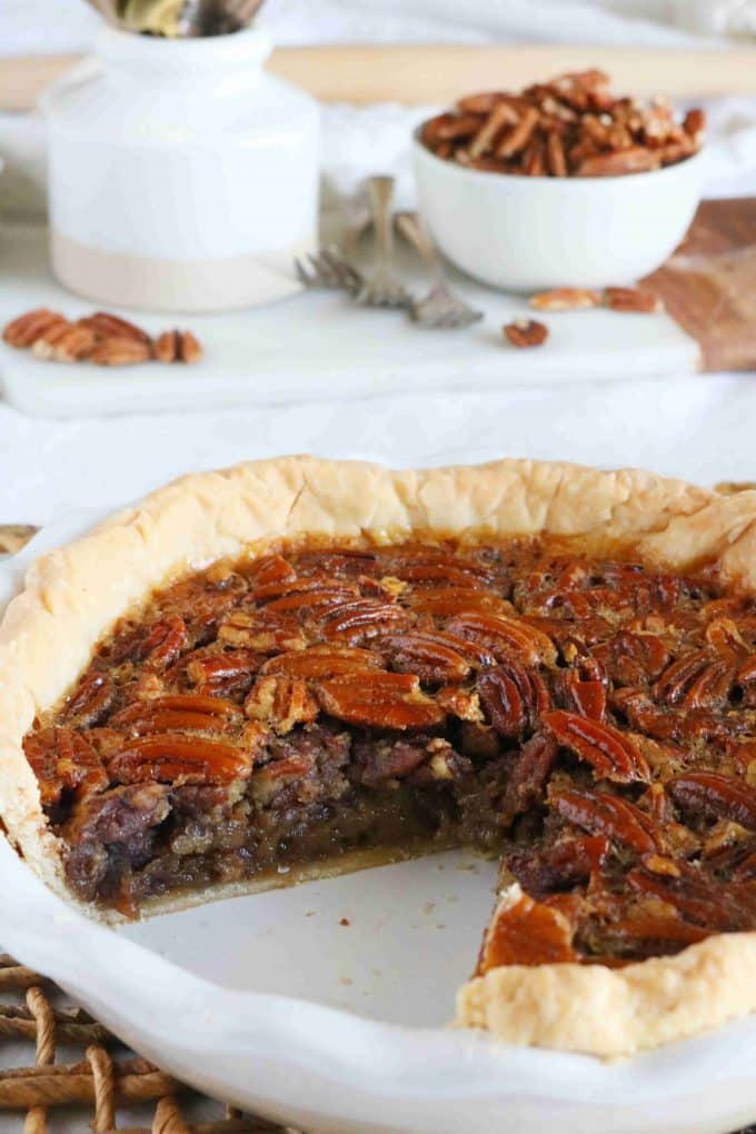 A close up shot of pecan pie in a white pie dish with a slice of pie missing. In the background is a white bowl full of whole pecans. 