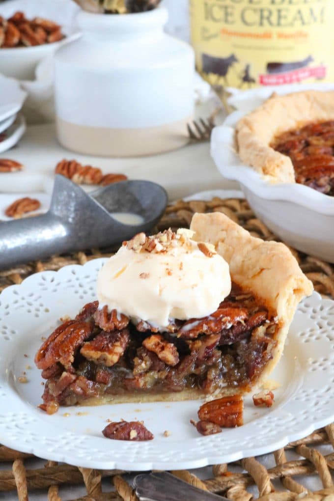 A slice of pecan pie topped with vanilla ice cream served on a white plate. In the background is an ice cream scoop, a gallon of ice cream and the whole pie. 