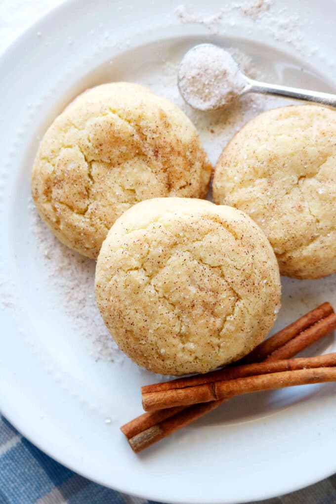 An overhead shot of three snickerdoodle cookies along with a spoon full of cinnamon sugar and two cinnamon sticks. 