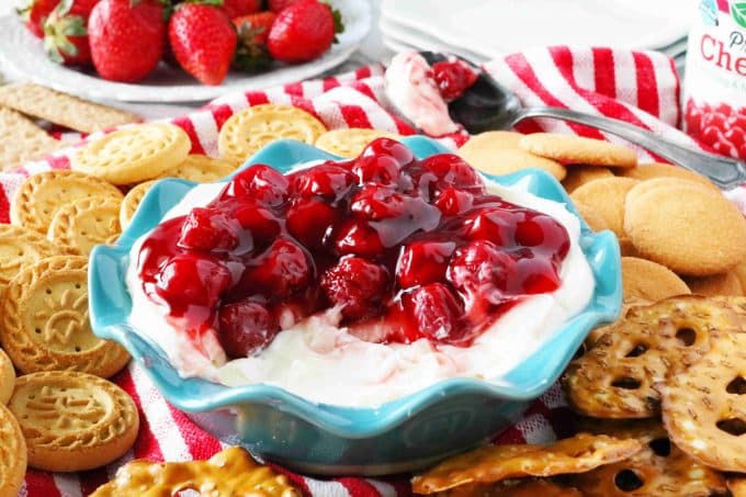 Cherry cheesecake dip served in a teal scalloped bowl surrounded by round cookies and pretzel thins. In the background is a plate of whole strawberries. 