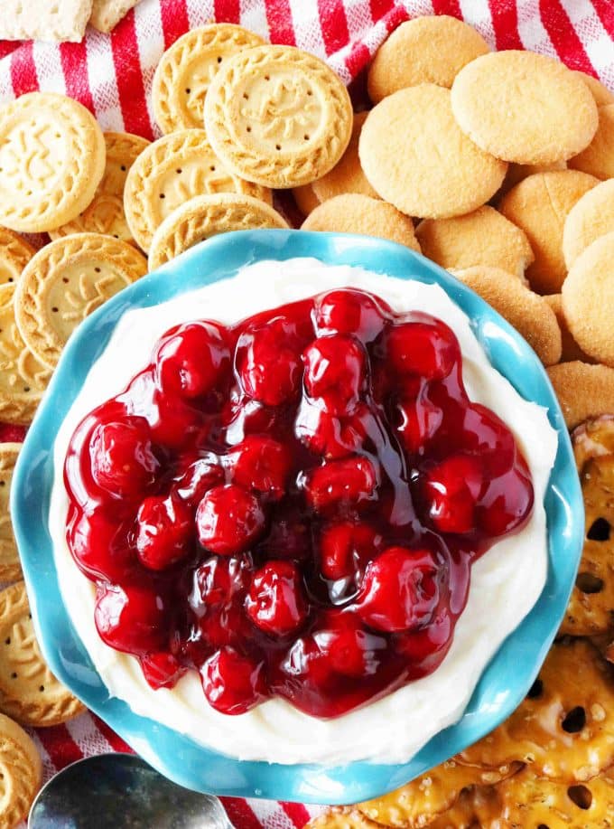 An overhead shot of cherry cheesecake dip served in a teal scalloped bowl surrounded by round cookies and pretzel thins. 