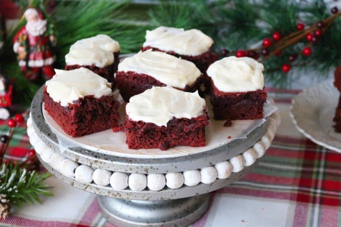 Red velvet brownies squares topped with cream cheese frosting served on a white pedestal stand. In the background are Christmas greenery and a Santa figurine. 
