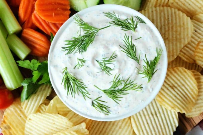 An overhead shot of dill dip surrounded by potato chips, carrots, celery sticks, and cucumbers. 