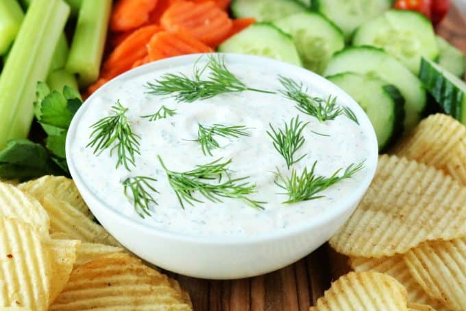 Dill Dip that is in a white bowl surrounded by potato chips, cucumbers, carrots and celery sticks. 