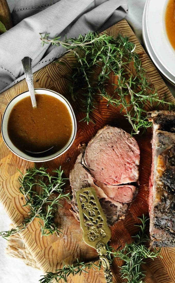 An overhead shot of Prime Rib Au Jus in a white bowl sitting next to a slice of prime rib. Fresh rosemary sprigs are placed around the beef and bowl sitting on a wood cutting board. 