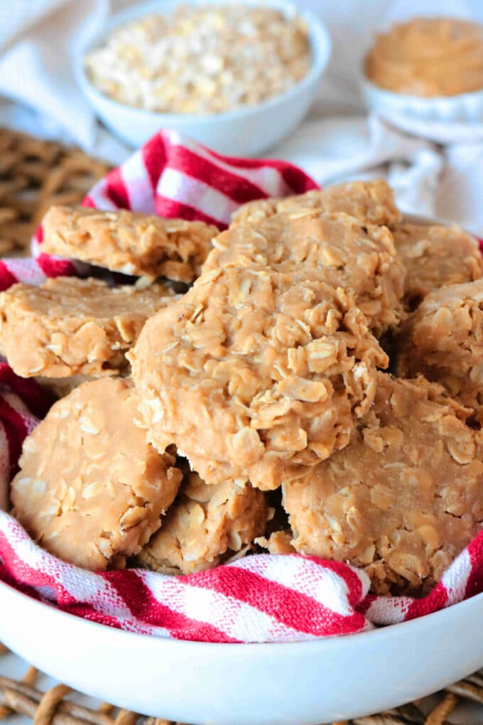 A white bowl with a red and white checkered napkin with Peanut Butter No Bake Cookies sitting on top. In the background are a bowl of oats and peanut butter. 