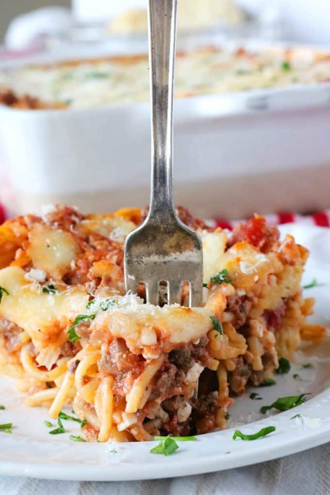 A triangle piece of baked spaghetti casserole served on a white plate. In the middle of the piece is a silver fork standing up. In the background is the white casserole dish. 