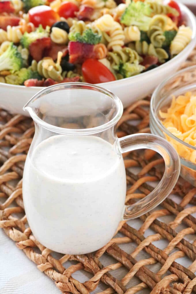 A small pitcher of ranch dressing with a bowl of the Bacon Ranch Pasta Salad behind it. Both items are sitting on top of a woven placemat. 