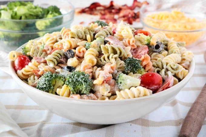 Bacon Ranch Pasta Salad served in a white bowl. In the background are smaller individual bowls of broccoli, bacon, and shredded cheese. 