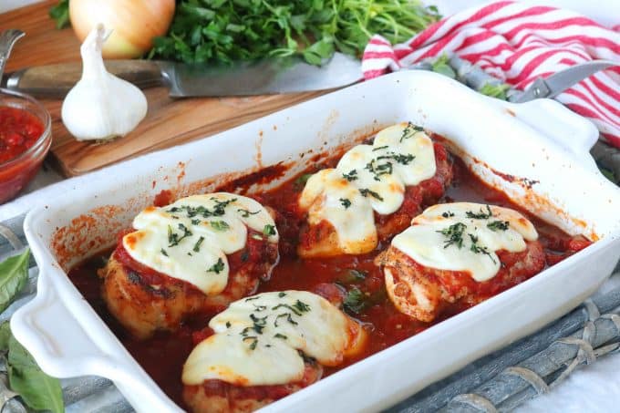 Four chicken breasts topped with mozzarella and marinara sauce that have been baked in a white casserole dish. Behind the dish is are the ingredients used to make the dish sitting on a wood cutting board along with a knife. 