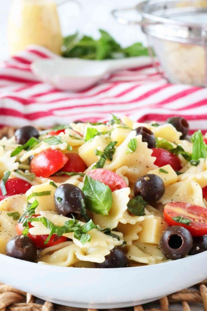 A close up shot of a white bowl of Bowtie Pasta Salad. Behind the bowl of pasta is a red and white checkered napkin, a white spoon, a pitcher of dressing, fresh basil leaves and a colander. 