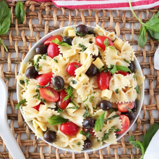 An overhead shot of a bowl of Bowtie Pasta Salad sitting on top of a wooden woven placemat. Sprinkled around the bowl are fresh basil leaves. 