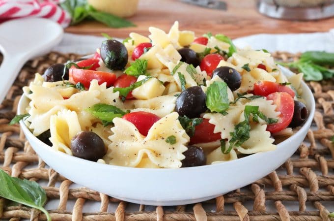 Bowtie Pasta Salad served in a white bowl sitting on top of a woven placemat. The salad features black olives, cherry tomatoes, fresh basil leaves and bowtie pasta. 