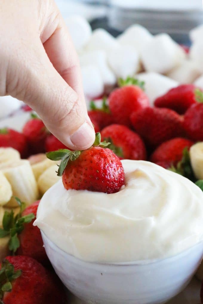 A hand is dipping a whole strawberry into the bowl of Fruit Dip. Surrounding the dip are strawberries, bananas and marshmallows. 
