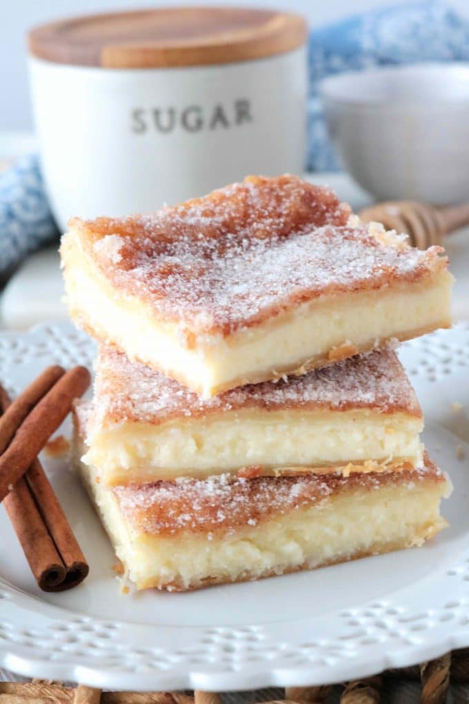 Sopapilla Cheesecake squares stacked up on top of each other on a white plate. Lying next to the squares are two cinnamon sticks. 