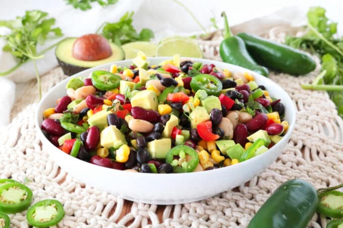 Mexican Bean Salad served in a white bowl surrounded by avocado halves, whole and sliced jalapenos. 