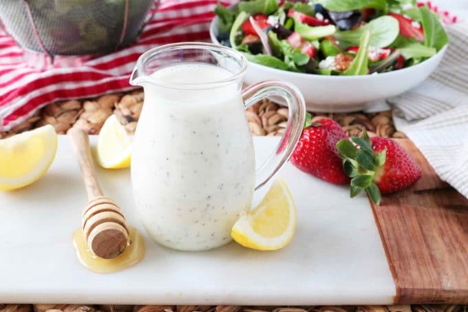 Poppy Seed Dressing served in a small glass pitcher. Lying next to the dressing is a honey spoon, lemon wedges and whole strawberries. Behind the pitcher of dressing a bowl of salad with fresh berries. 